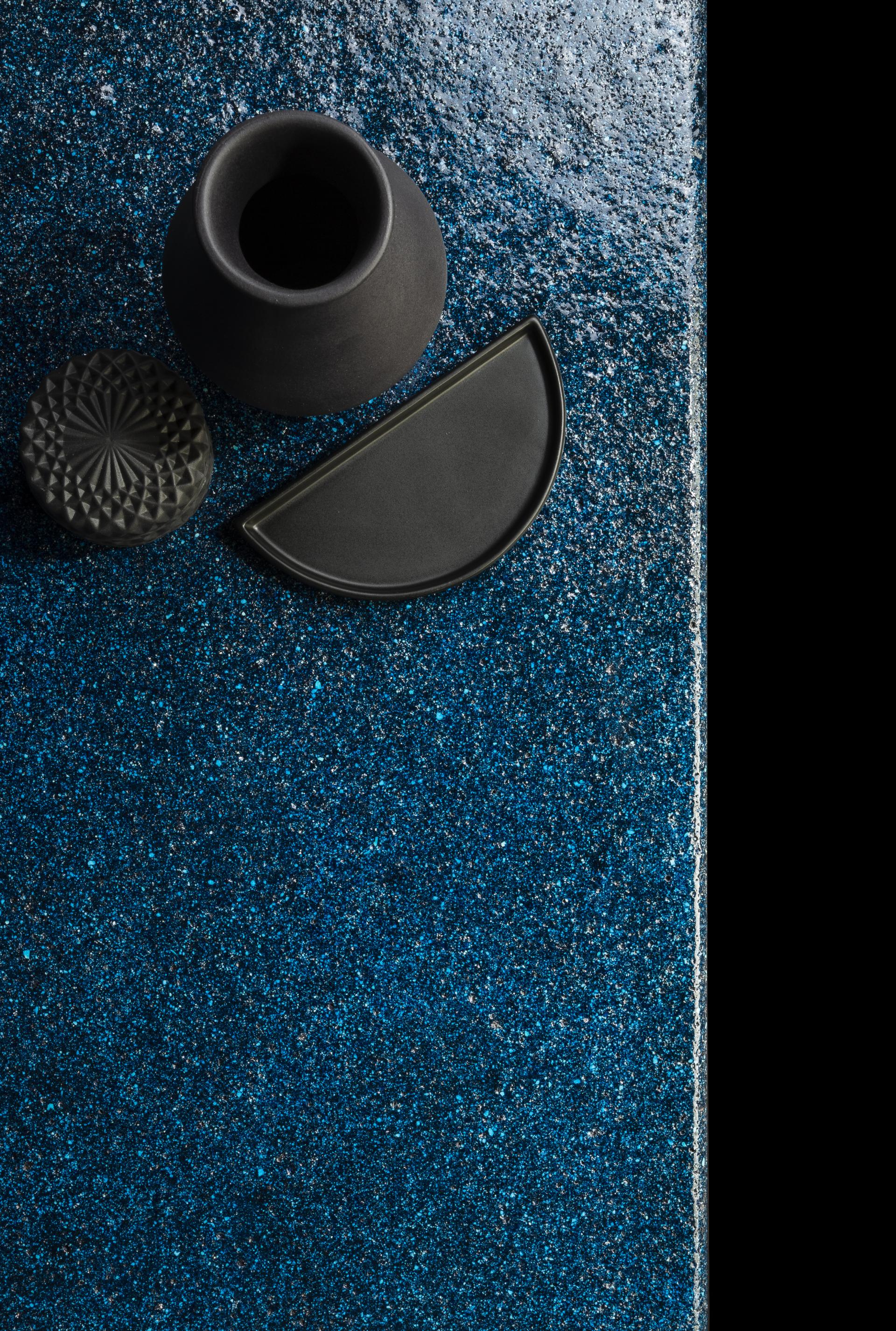 Black ceramic vessel, geometric plate, and textured lid on shimmering blue surface, highlighting design elements for Material Source Studio.