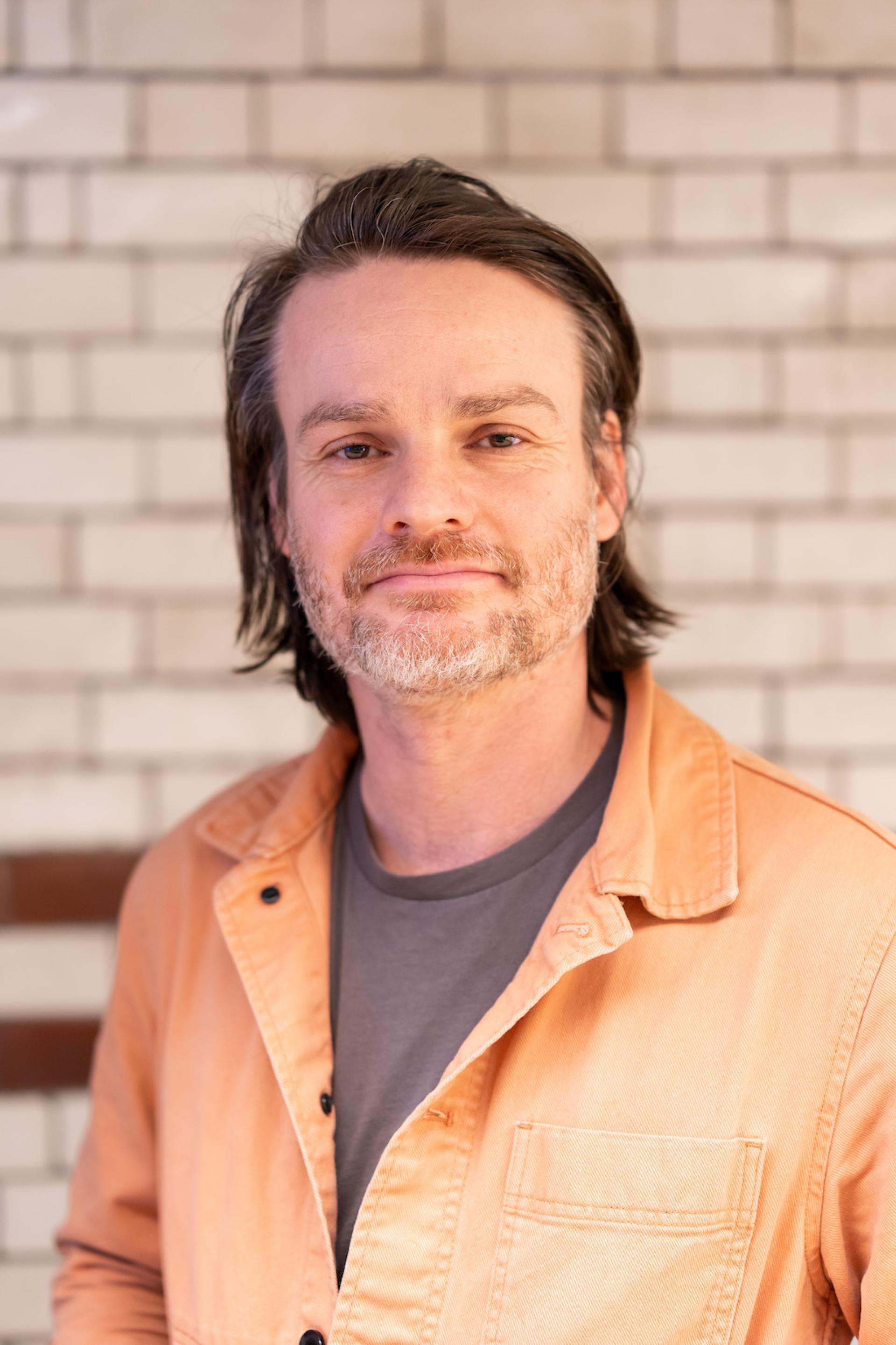 Man in an orange work jacket smiles in front of a tiled background, reflecting themes of sustainability and value engineering.