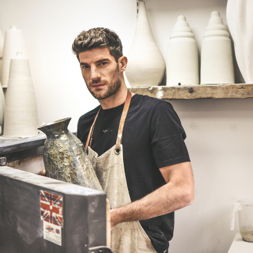 Ceramicist Ryan Barrett holding a handcrafted vase in his studio, surrounded by artistic pottery and tools.
