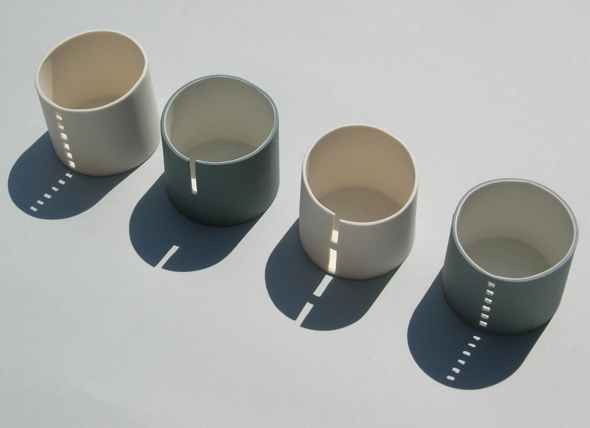 Four ceramic cups in various colors display unique cut-out designs, casting interesting shadows on a light background.