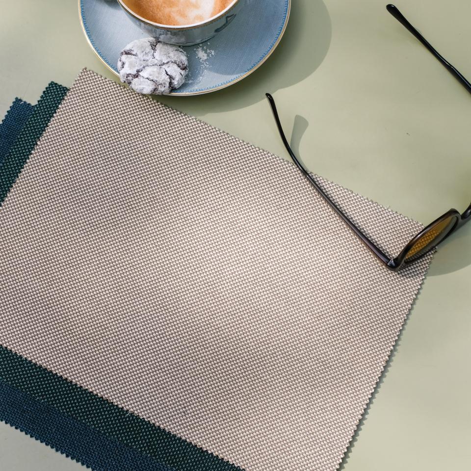 Two-tone woven fabric swatches from Agua's Zephyr collection alongside a cup of coffee and sunglasses on a green table.