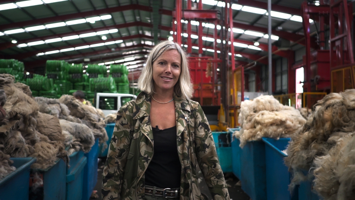 Award-winning designer Ella Doran stands in a vibrant wool-processing facility surrounded by bales of British wool.
