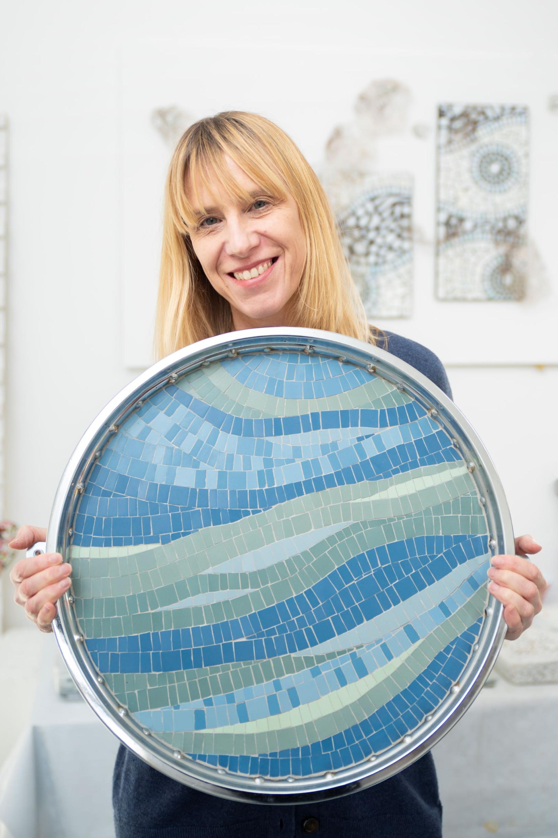 Craft artist proudly displays a vibrant mosaic artwork featuring blue and green wave patterns in Nottingham studio.