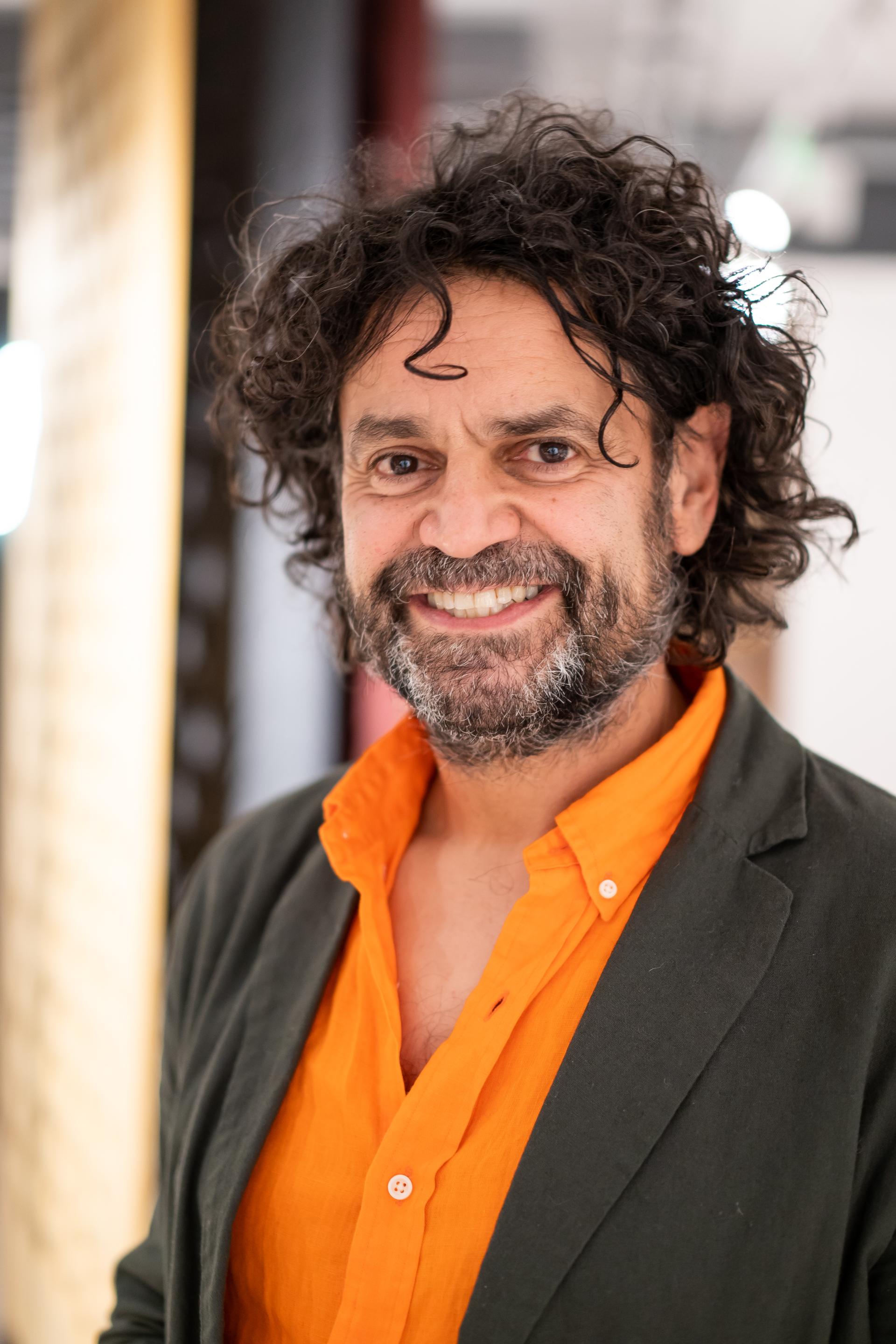 Smiling man with curly hair wearing an orange shirt and dark blazer, discussing sustainability at a seminar.