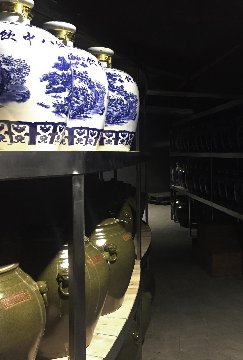 Ceramic jars with blue designs and olive-green pots on shelves in a dimly lit Chinese liquor factory.