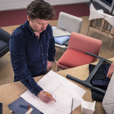 Design director Nick Lewis sketches furniture concepts amidst colorful fabric samples and modern chairs in a workshop.