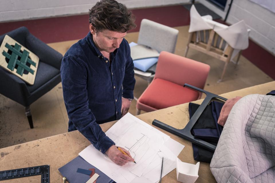 Design director Nick Lewis sketches furniture concepts amidst colorful fabric samples and modern chairs in a workshop.