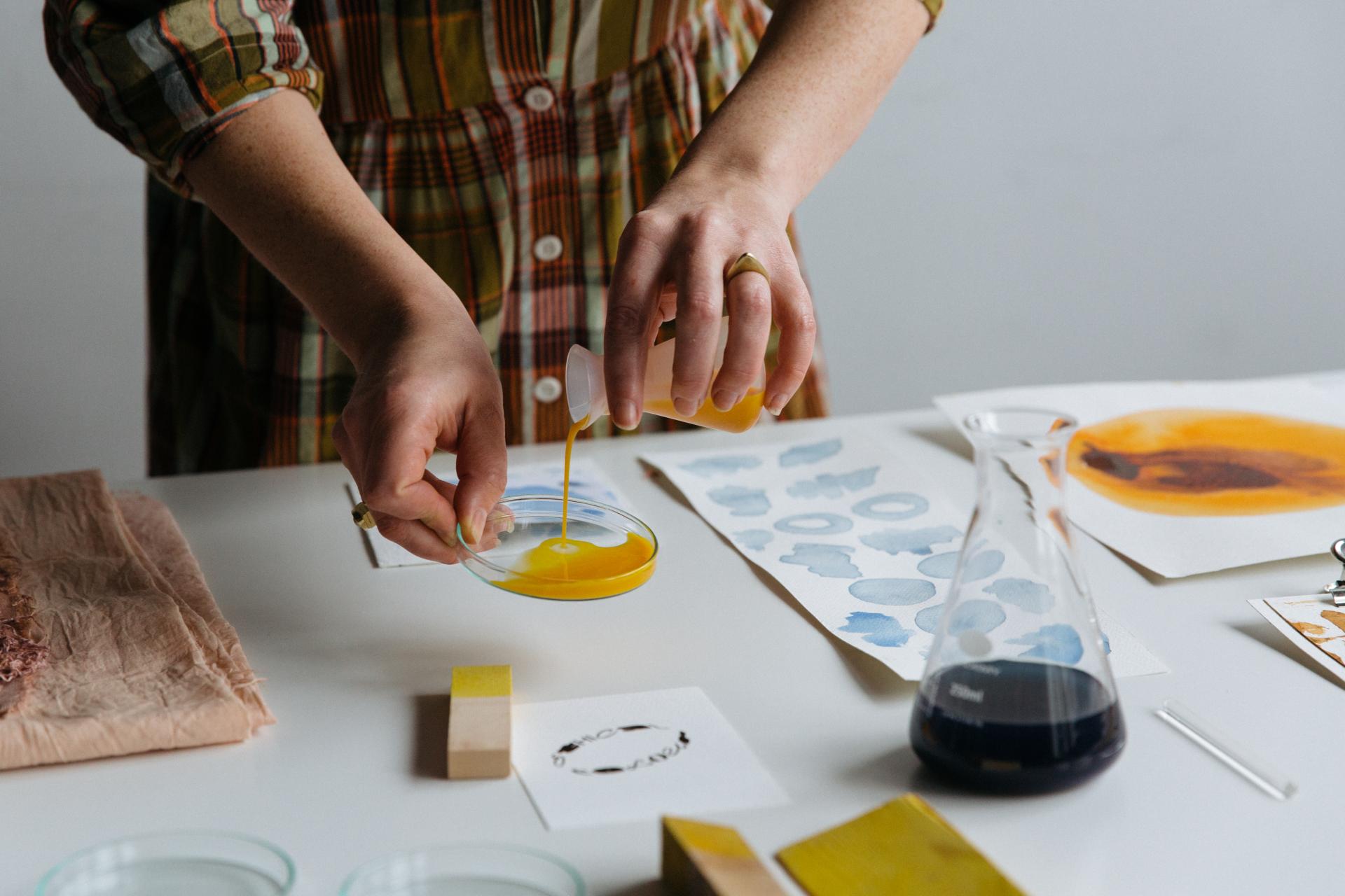 Hands pouring yellow liquid into a petri dish, surrounded by sketches and materials related to color design and sustainability.