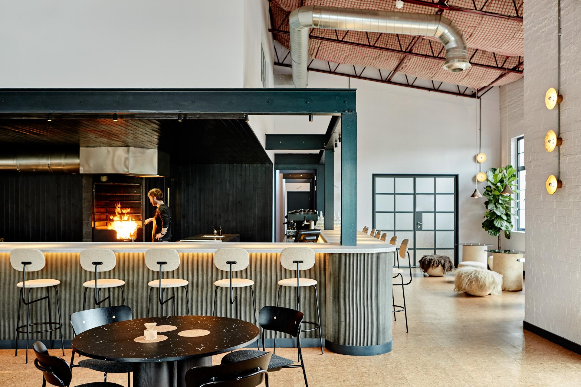 Contemporary restaurant interior featuring a bar, seating, and a chef cooking over an open flame, highlighting sustainable design.