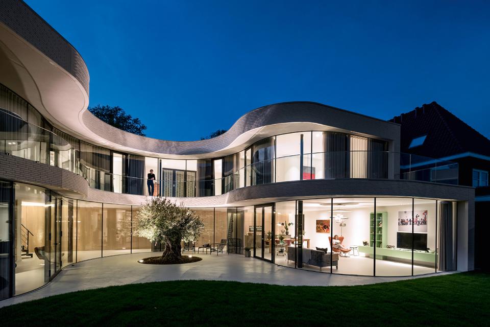 Modern architectural masterpiece featuring glass walls, a central olive tree, and inviting outdoor spaces at dusk.