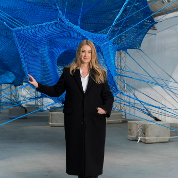Anya Hindmarch stands in her immersive Weave Project installation, surrounded by vibrant blue netting and structural elements.