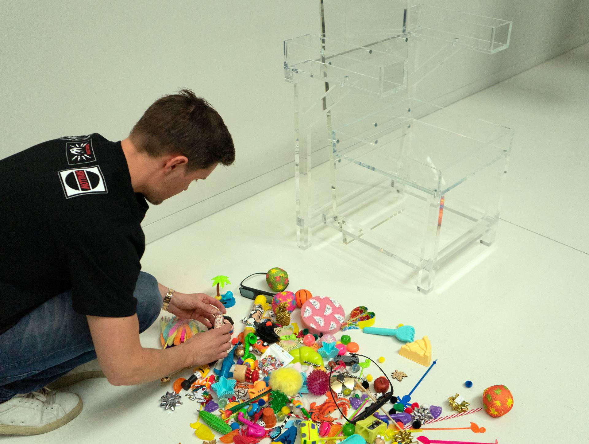 Ruud van Eijk selecting colorful objects from a pile, with a transparent chair model in the background, showcasing creativity and design.