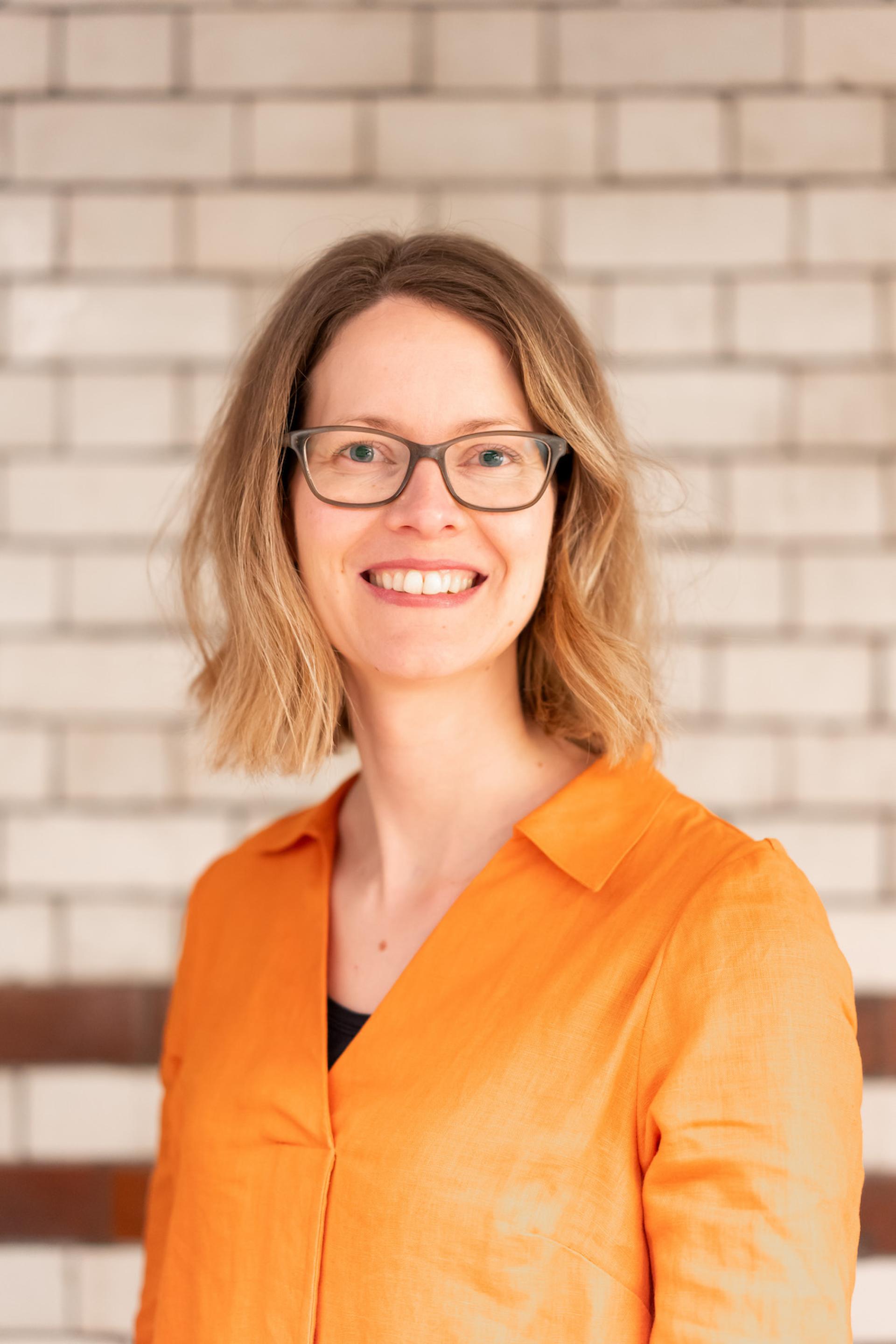 Smiling woman in an orange shirt, standing against a brick wall, representing discussions on sustainability and value engineering.