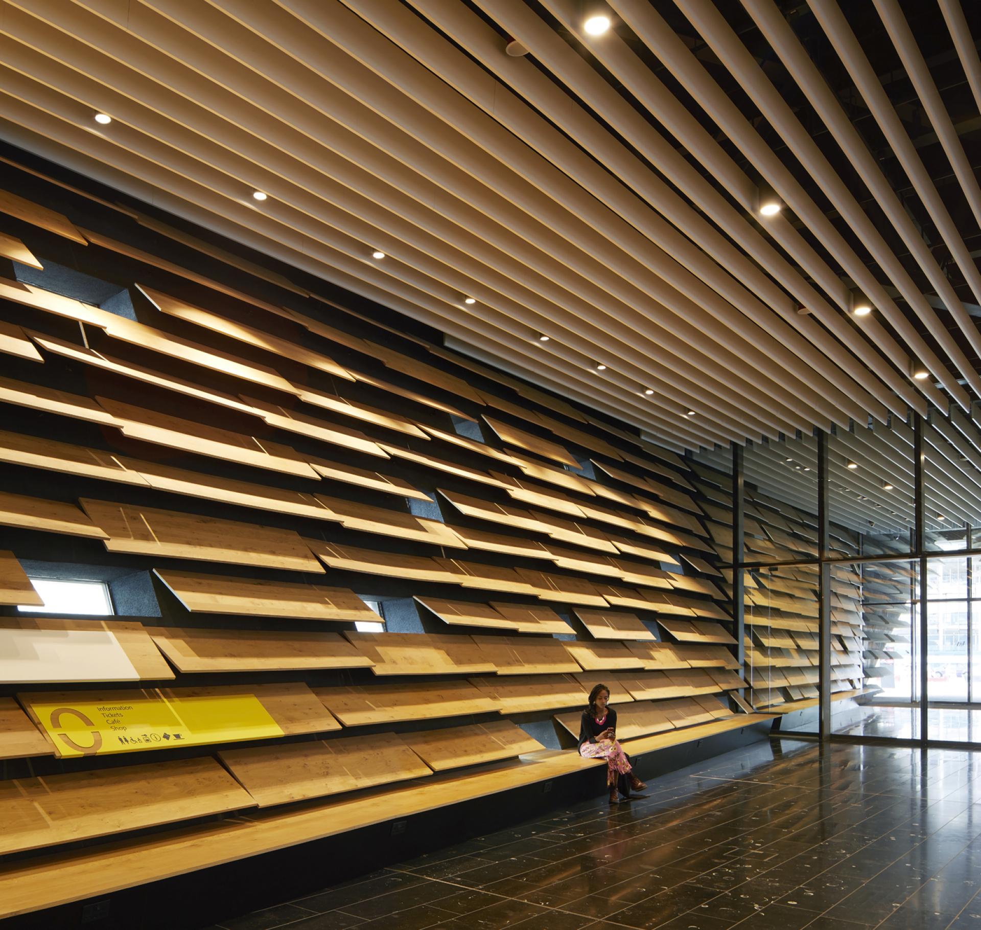 Bright interior of V&A Dundee showcasing wooden wall features designed by Kengo Kuma, with a visitor seated nearby.