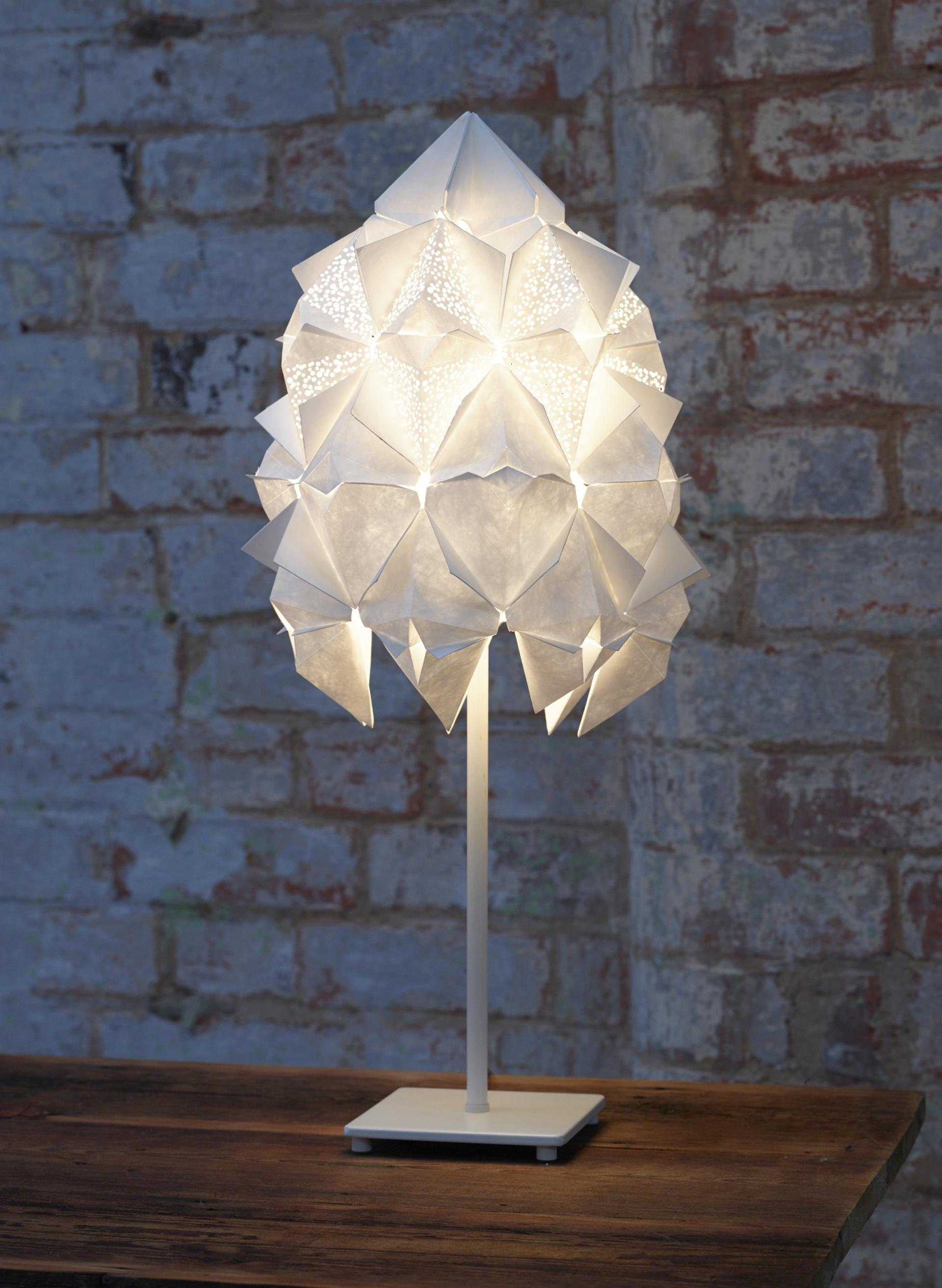 Sculptural white lamp with geometric design illuminating a rustic wooden table against a textured brick wall.