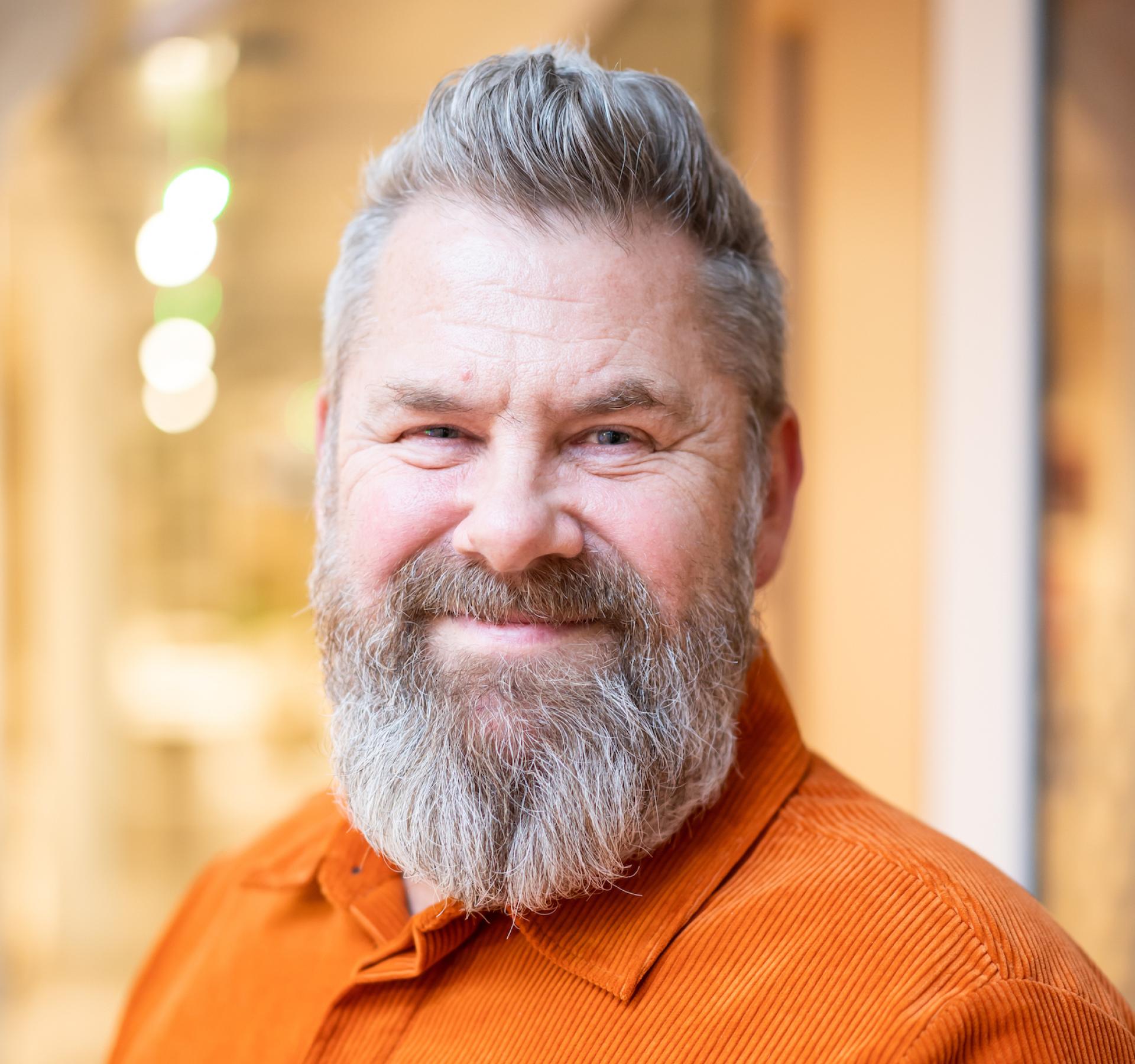 Smiling educator with a beard wearing an orange shirt, representing the evolving future of education and learning environments.
