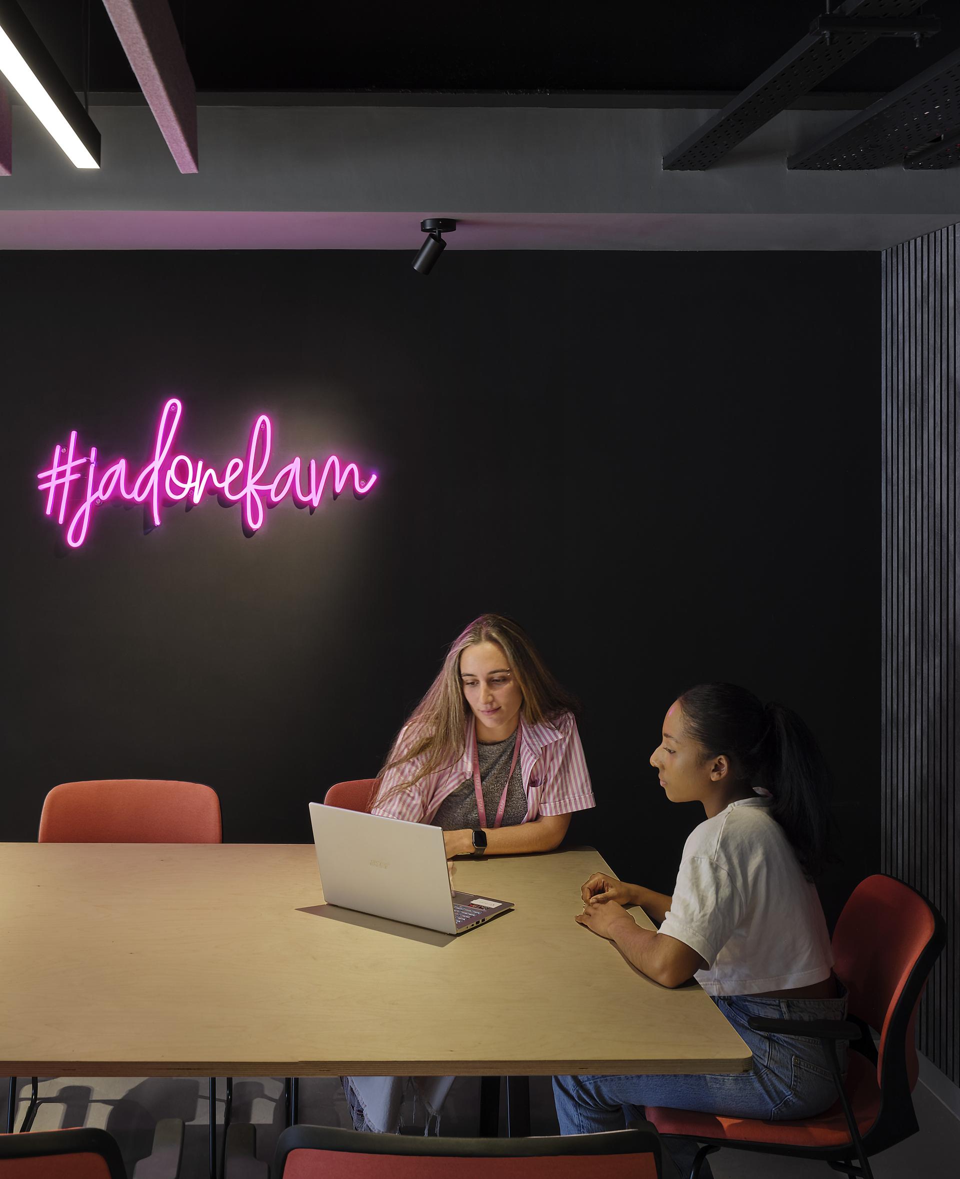 Two women collaborating at a modern table, with a neon "#jadorefam" sign illuminating a design studio.