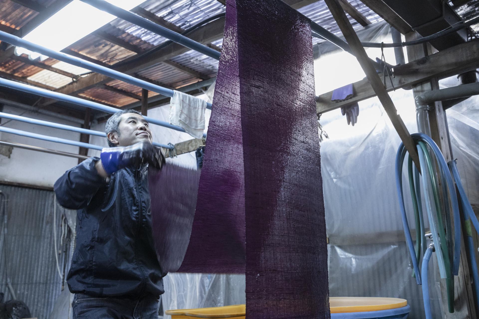 Artisan demonstrating traditional Japanese dyeing techniques with vibrant purple fabric in a workshop setting.