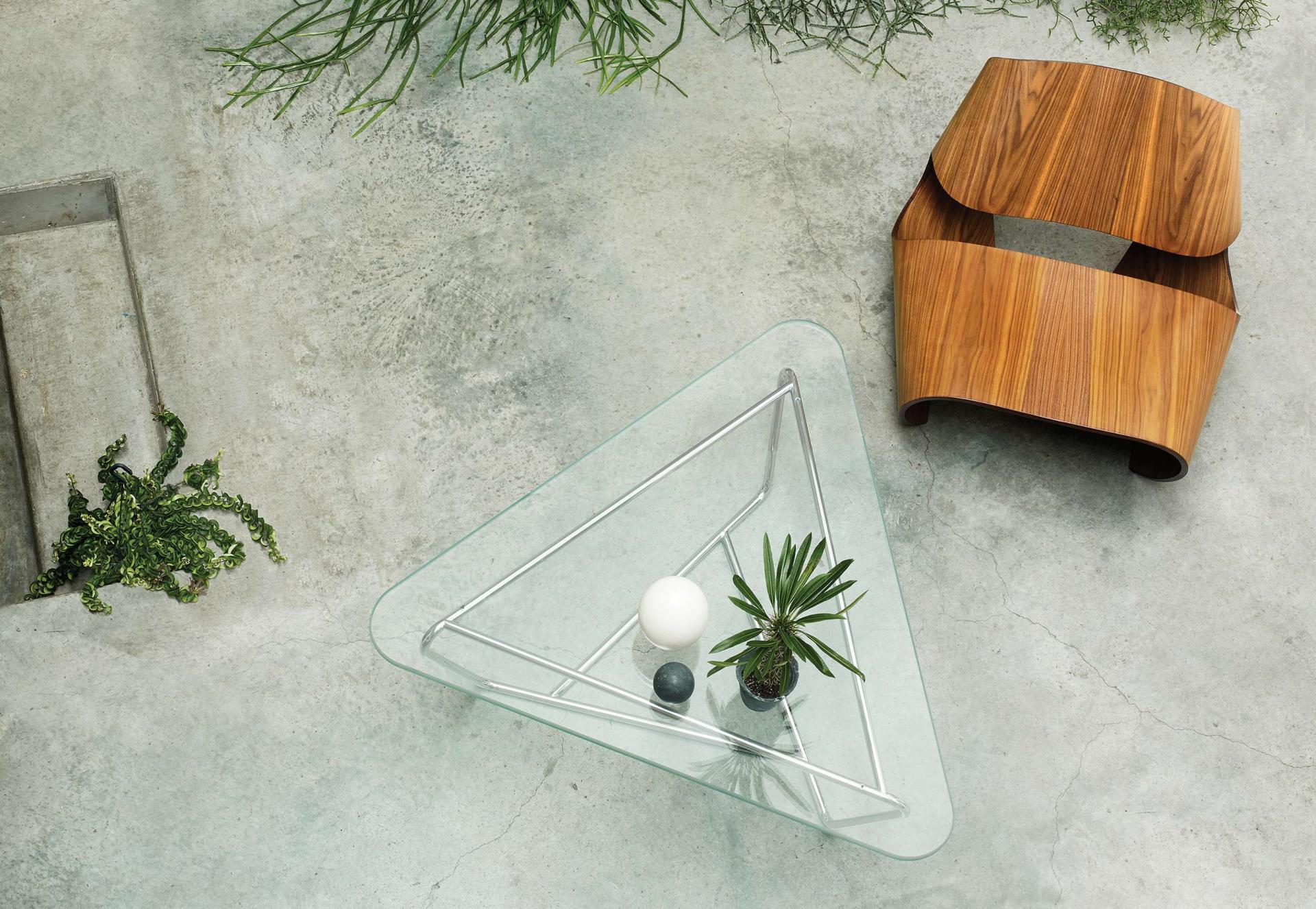 Modern triangular glass table with decorative plants next to a curved wooden chair on a textured concrete floor.