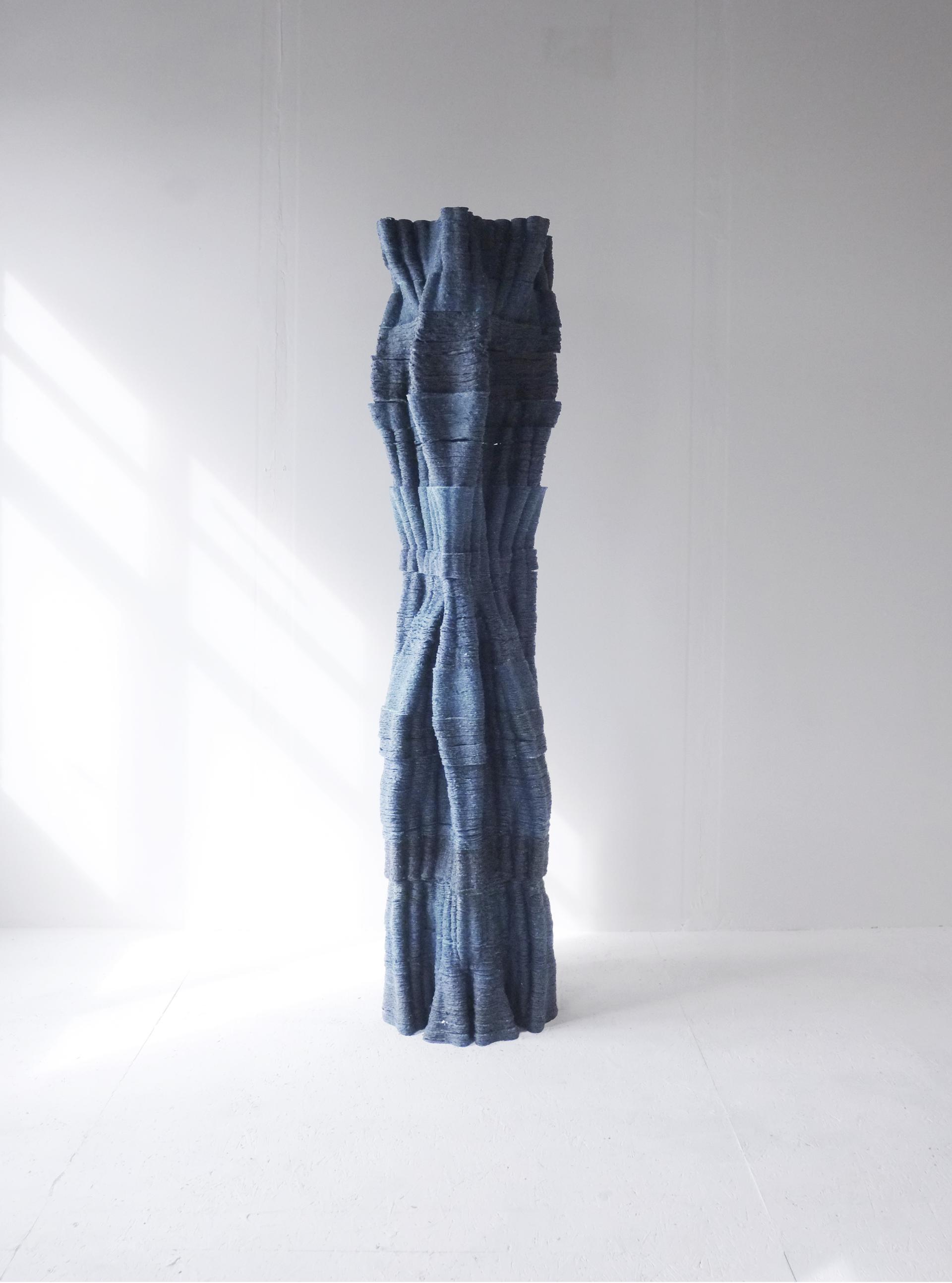Sculptural representation of a tree trunk, showcasing textured blue fibers in a minimalist studio setting.