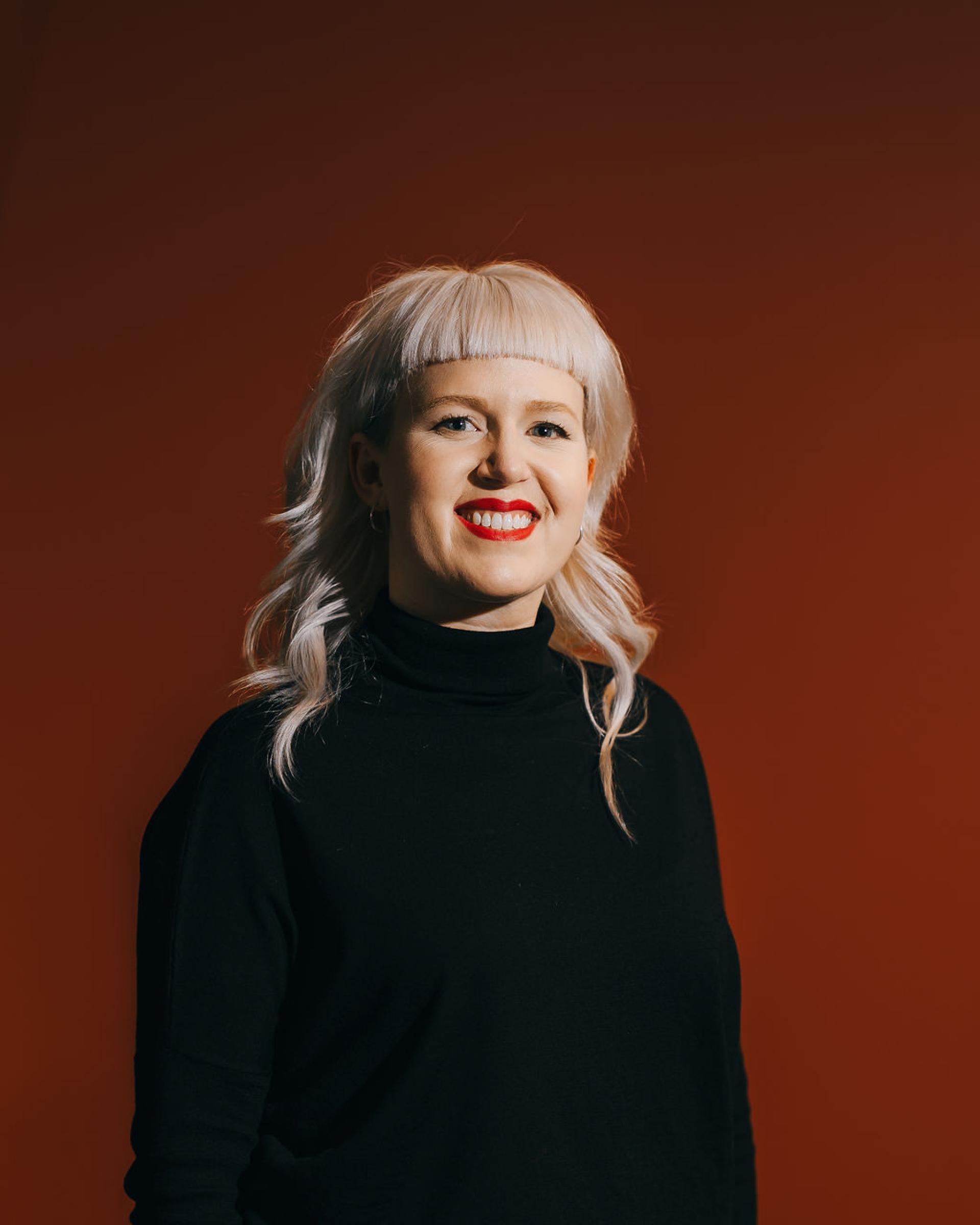Smiling woman with blonde hair in a black turtleneck, against a warm, red background, promoting sustainability in biomaterials.
