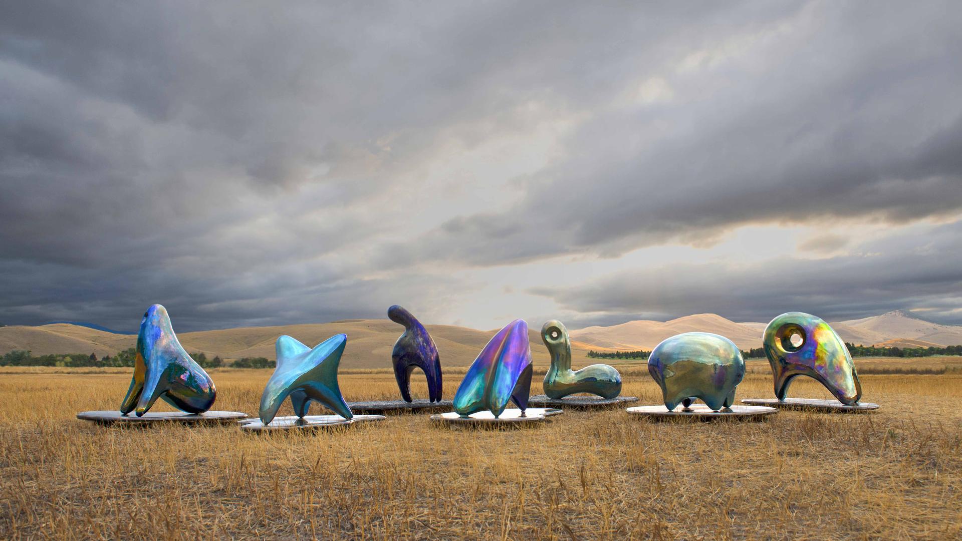 Colorful abstract sculptures displayed on pedestals against a dramatic sky, showcasing contemporary art in a natural setting.