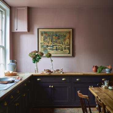 A stylish kitchen featuring Farrow & Ball's bold pink walls, dark cabinetry, and fresh flowers on the countertop.