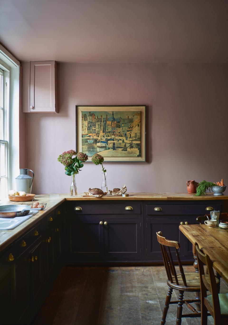 A stylish kitchen featuring Farrow & Ball's bold pink walls, dark cabinetry, and fresh flowers on the countertop.