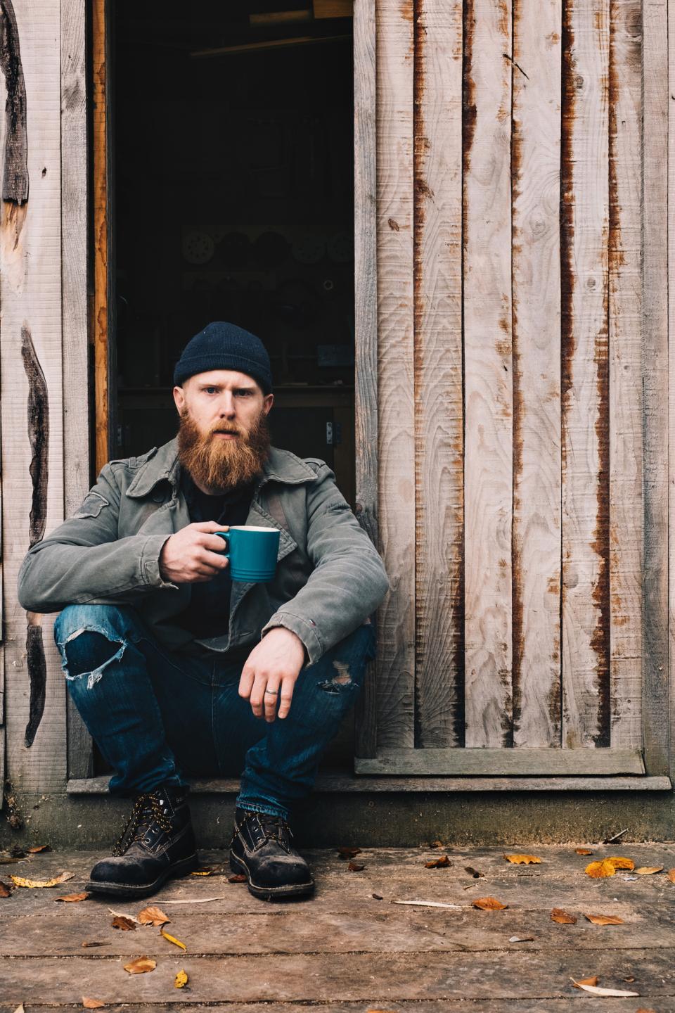 Pete Hill, a designer, sits on a doorstep, enjoying coffee, reflecting craftsmanship and local business.
