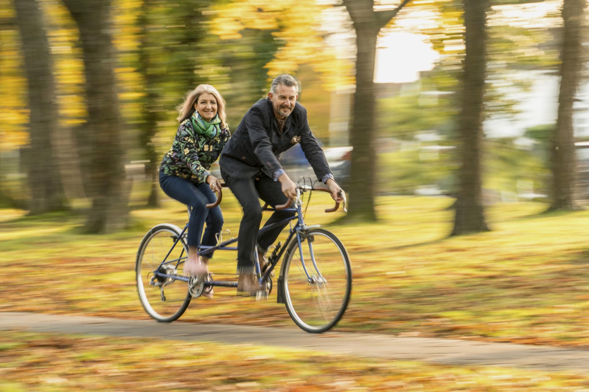 Couple riding a tandem bicycle through a vibrant park, surrounded by autumn leaves and trees, embracing joy and connection.