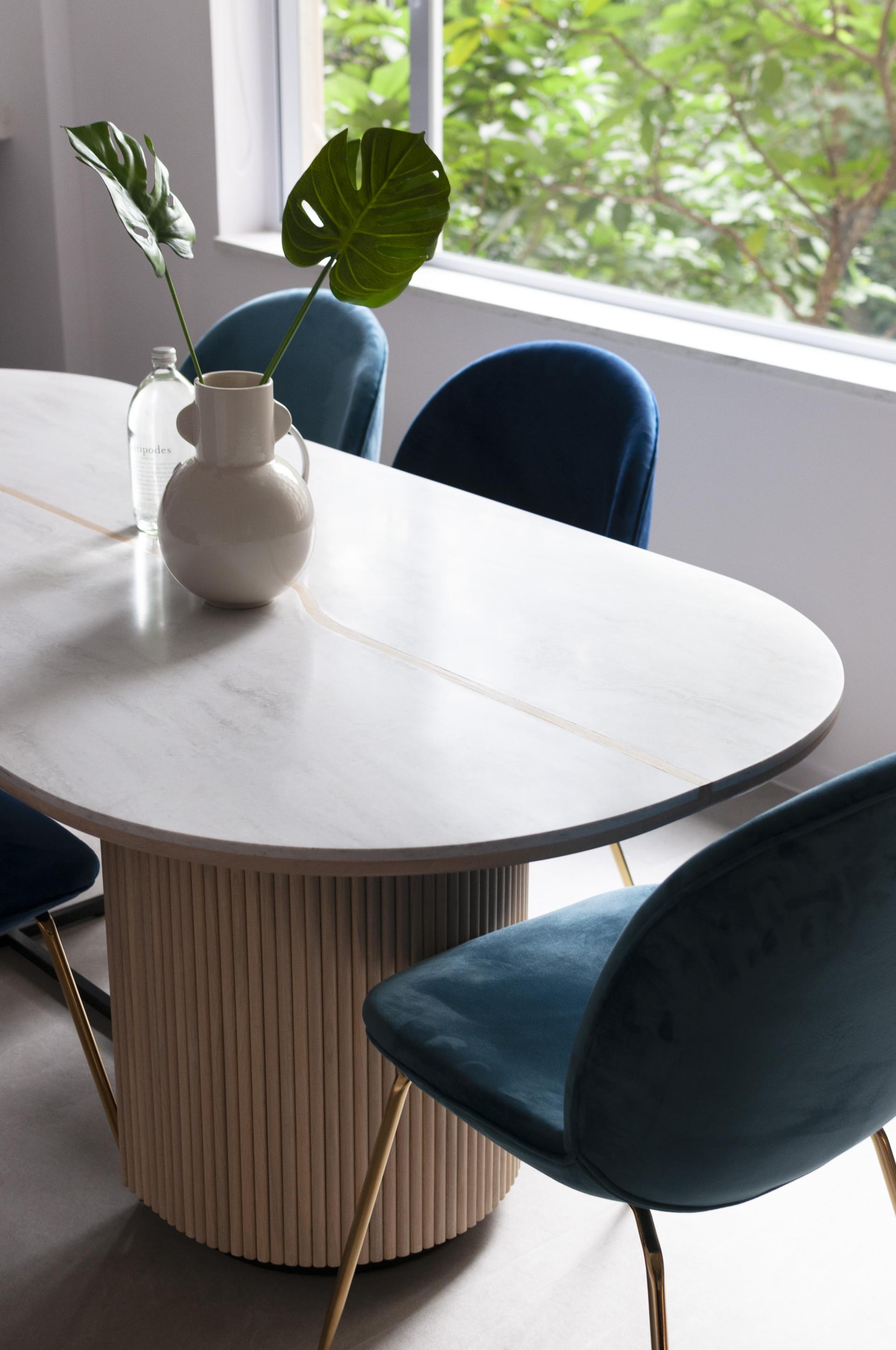 Stylish dining table with marble surface and modern blue chairs, featuring tropical leaves in a vase by the window.