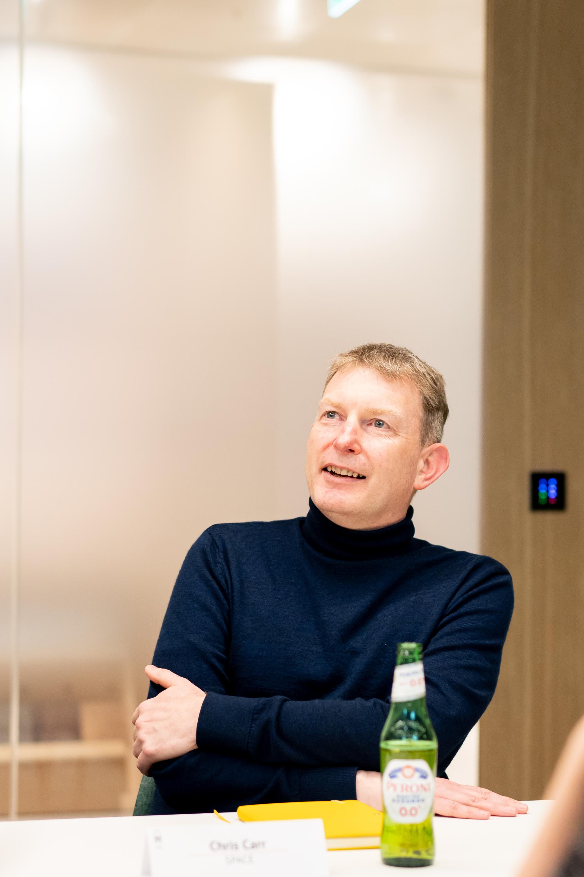 Man in a turtleneck sweater discussing workplace flexibility, with a drink and notepad on the table.