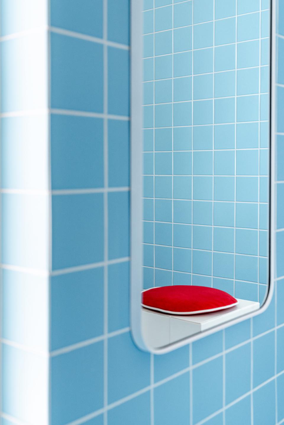 Bright blue tiled wall with a reflective mirror framing a red cushion, evoking a Wes Anderson-style aesthetic.