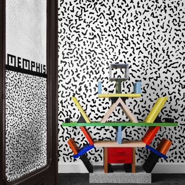 Colorful Memphis Milano furniture displayed against a playful black-and-white patterned wall in a modern gallery setting.