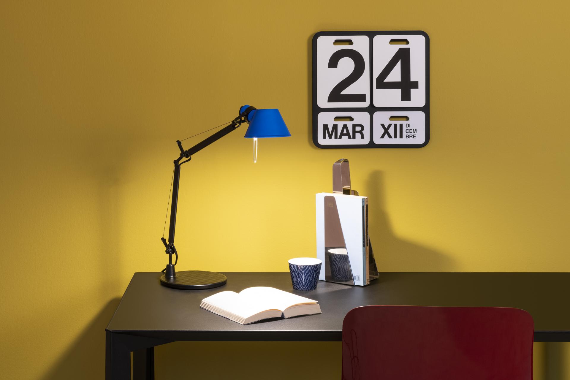 A stylish Tolomeo Micro lamp illuminates a modern workspace featuring books, a cup, and a calendar against a vibrant yellow wall.