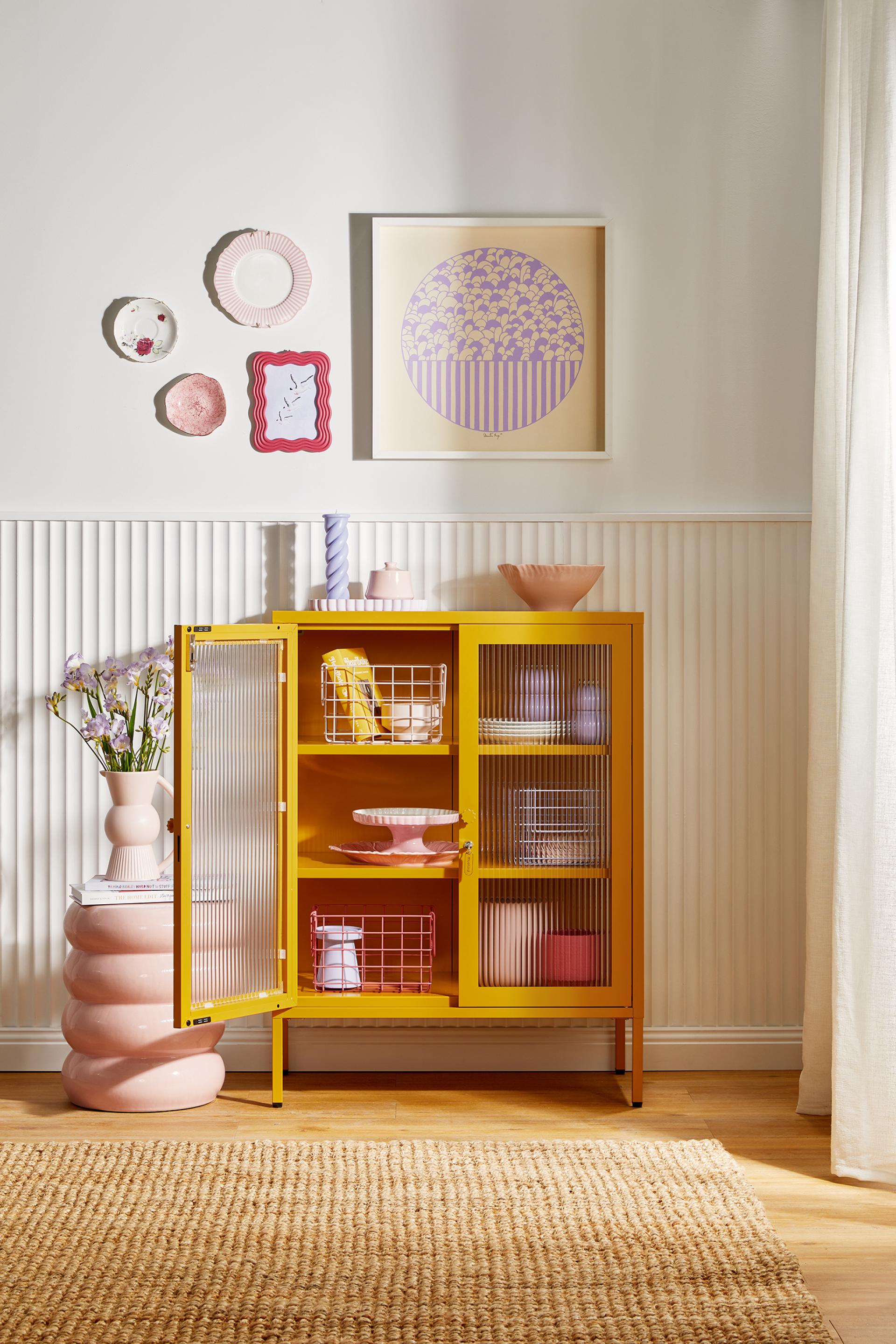 Vibrant yellow cabinet from Mustard Made's Glass Collection, featuring decorative tableware and artwork on a light wall.