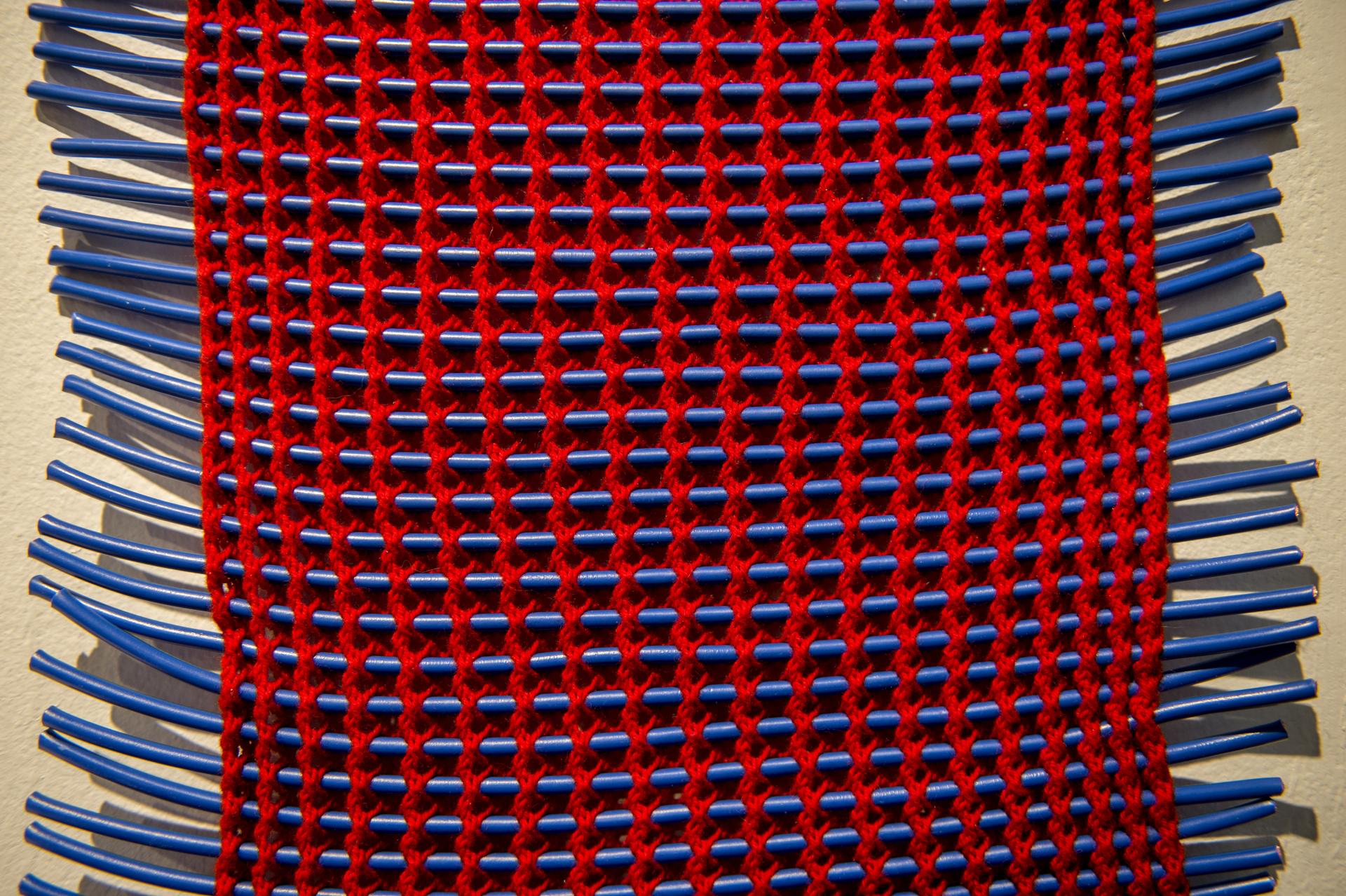 Vibrant woven textile artwork featuring red yarn and blue tubes, showcased at the Surface Design Show 2020 in London.