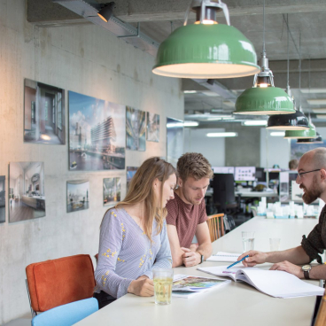 Three architects collaborate at a table in a modern studio, surrounded by architectural photography on the walls.