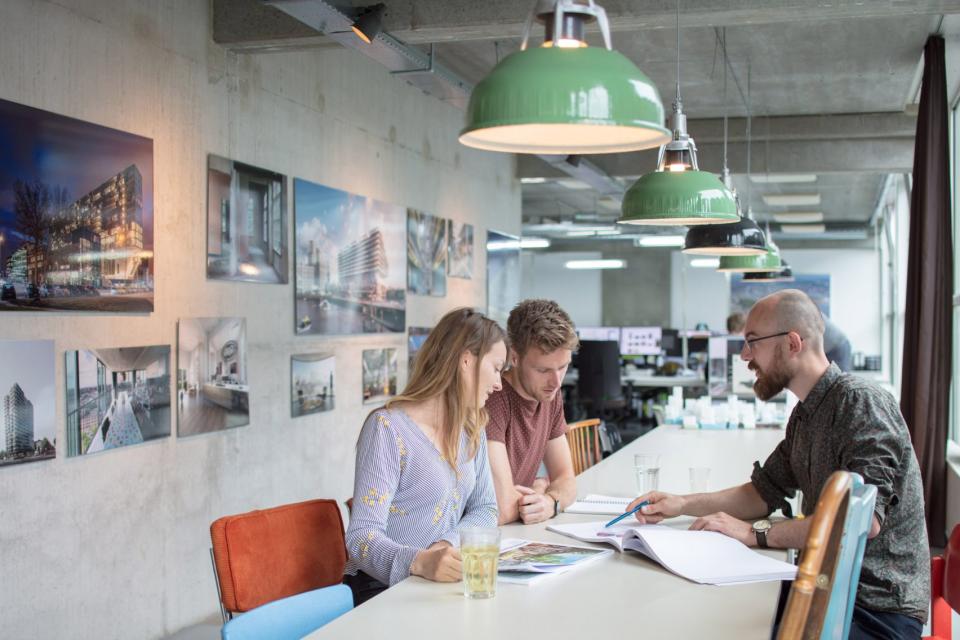 Three architects collaborate at a table in a modern studio, surrounded by architectural photography on the walls.
