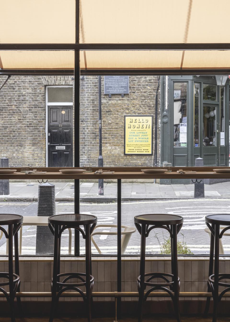 Interior view of Honey & Co Bloomsbury, featuring wooden bar stools and large windows showcasing the street outside.