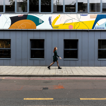 A woman walks along a street with vibrant, mood-boosting artwork on a building above her in a co-working area.