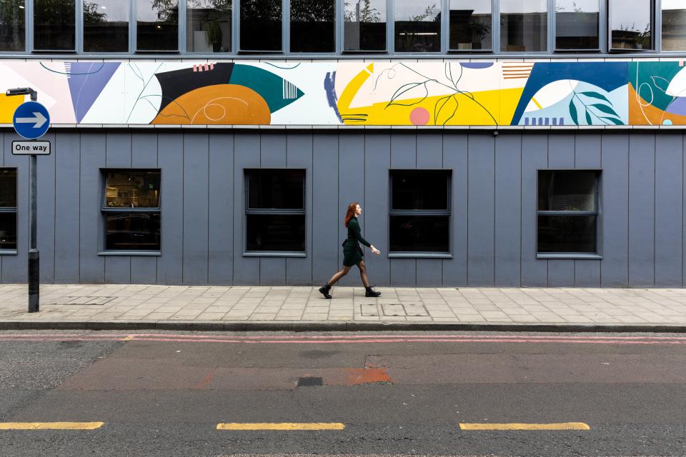 A woman walks along a street with vibrant, mood-boosting artwork on a building above her in a co-working area.