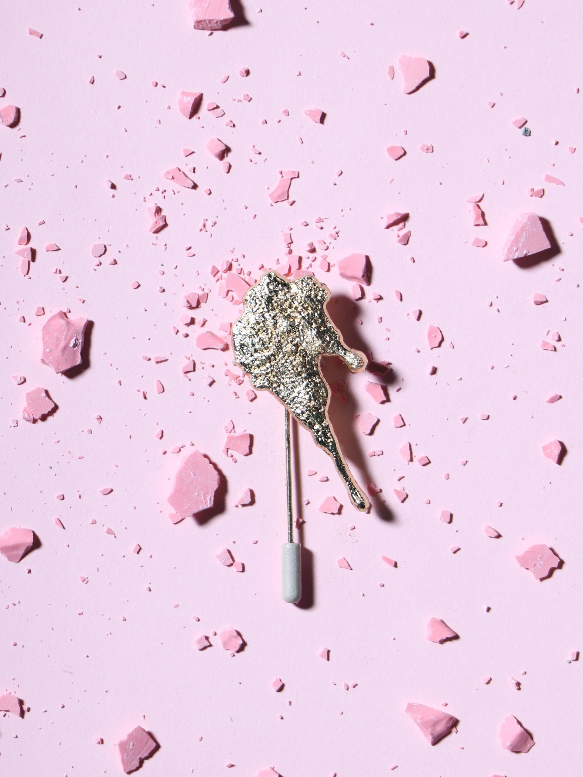 A shiny, sculptural brooch rests on a light pink surface scattered with pink shards, showcasing innovative design and material experimentation.