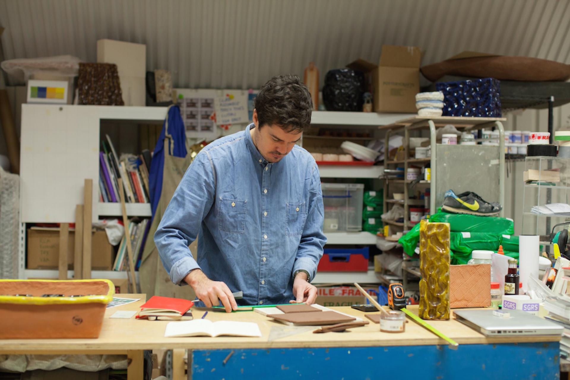Ceramic artist Matthew Raw works at his studio desk, surrounded by tools, clay, and artistic materials.