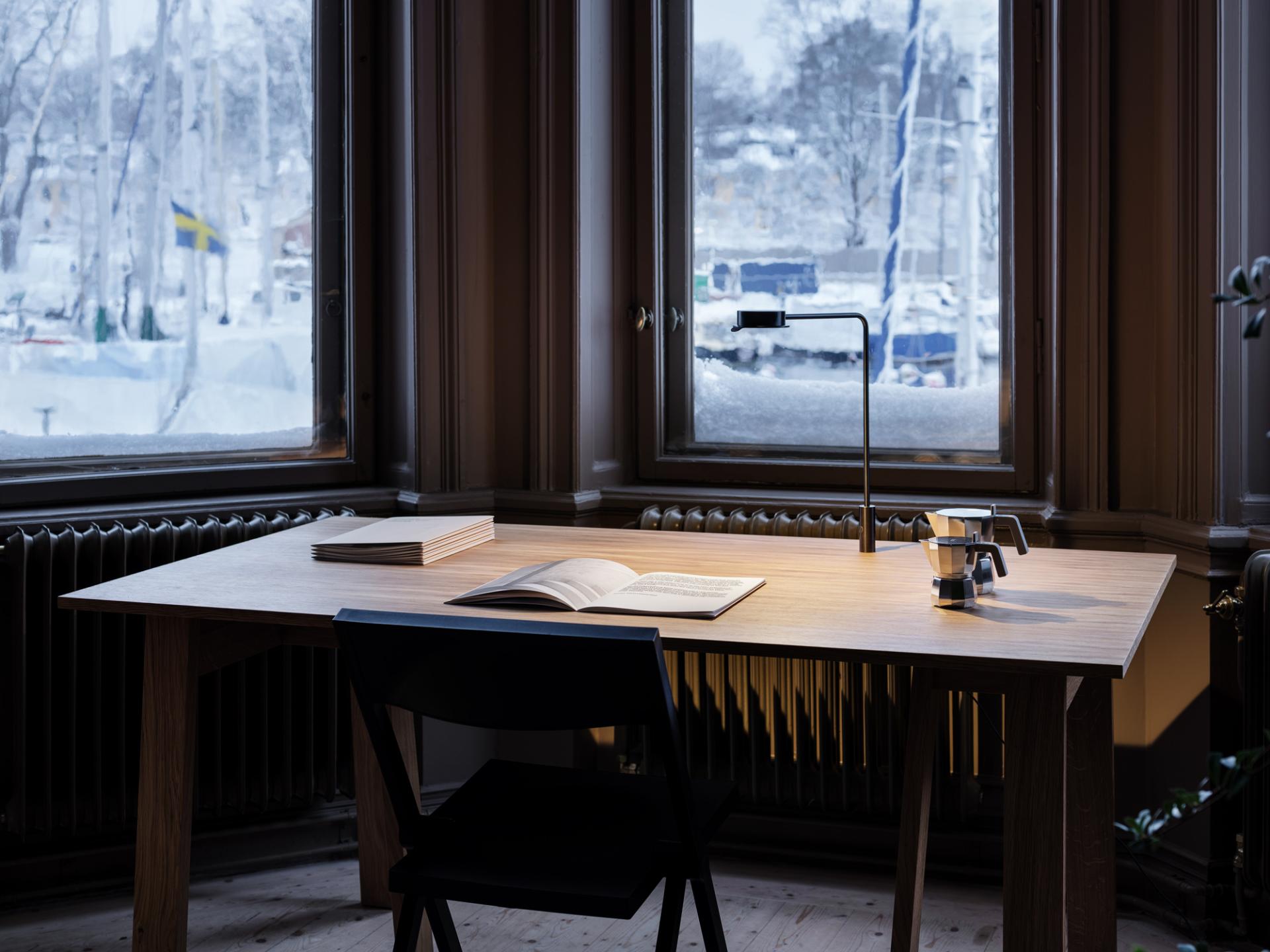 A modern w102 Chipperfield lamp illuminates a wooden desk with an open book and espresso maker by a snowy window.