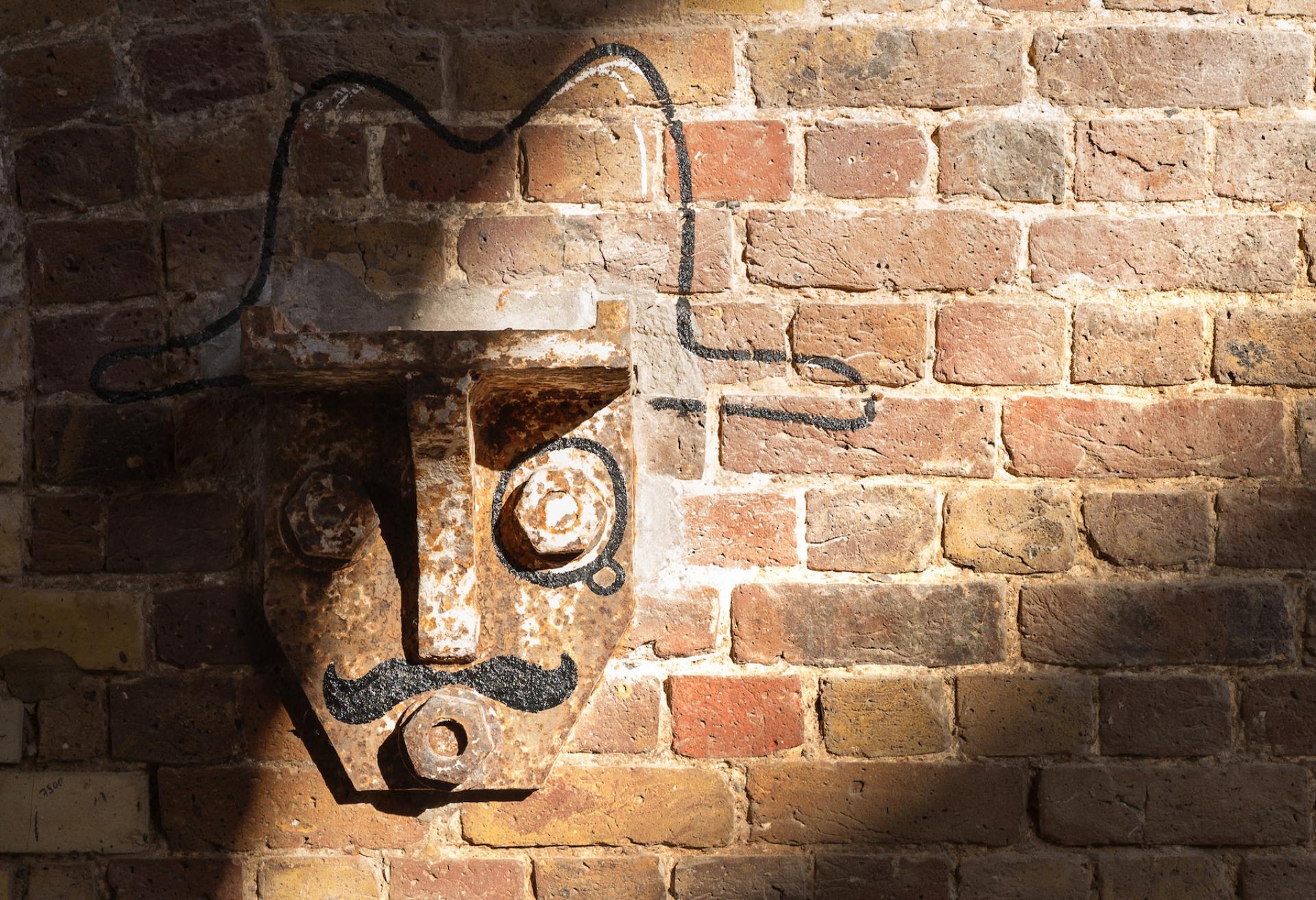 Rusty metal face sculpture with a mustache, creatively integrated into a brick wall at Borough Yards, London.