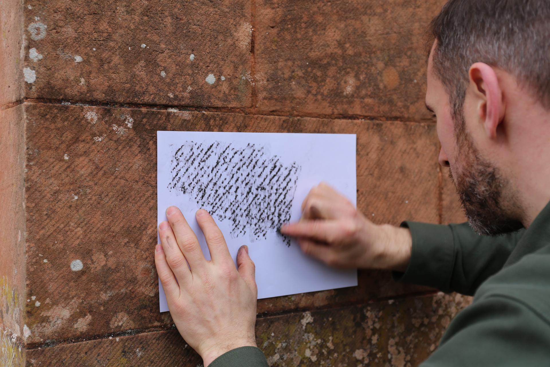 A man creating a textured rubbing on paper from a weathered stone wall, highlighting Bute Fabrics' artistry.