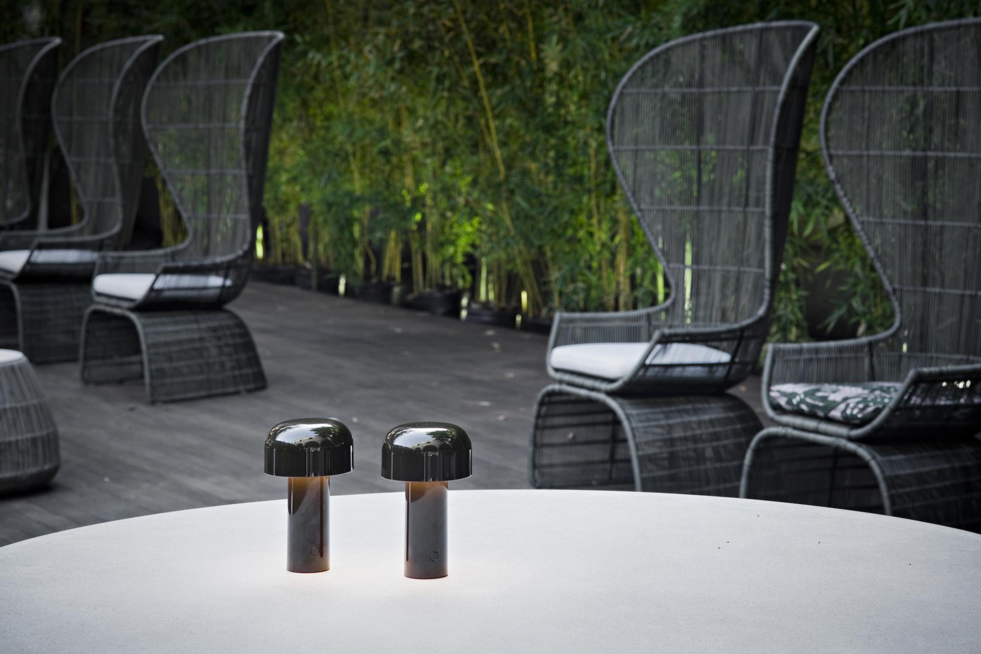 Portable outdoor lighting solutions by Flos on a modern table, surrounded by stylish woven chairs in a lush garden setting.