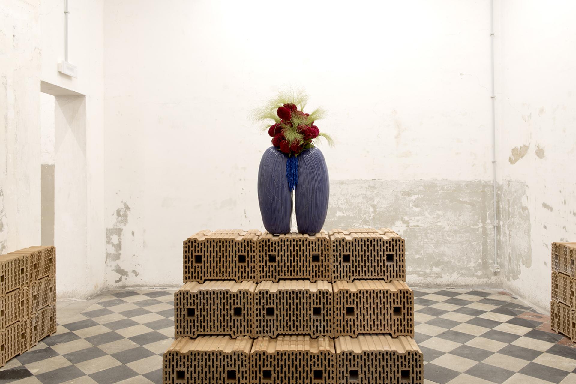 Unique vase with flowers displayed on a stack of textured bricks in a contemporary art space in Rome.