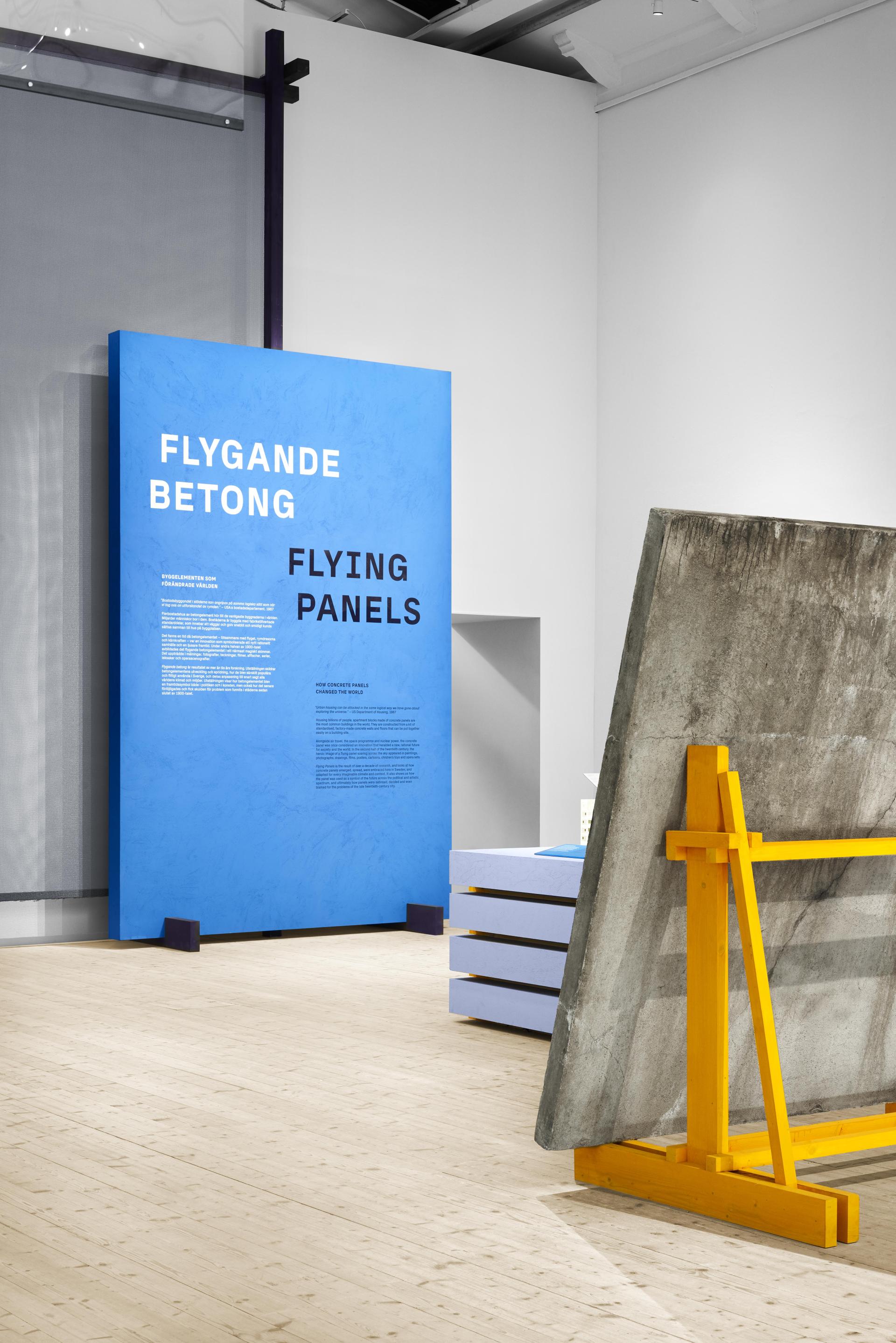 Exhibition display featuring a blue wall panel titled "Flying Panels," highlighting the impact of concrete in architecture.